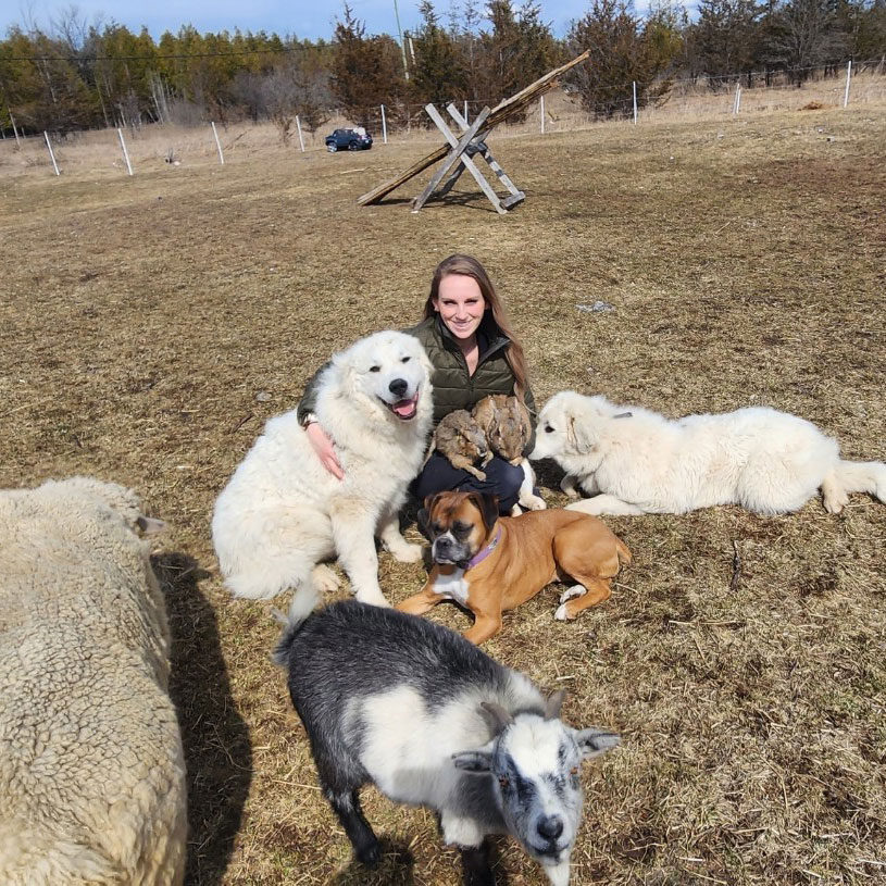 Reeva sits on the ground at her farm with three dogs, two bunnies, a sheep and a goat around her.